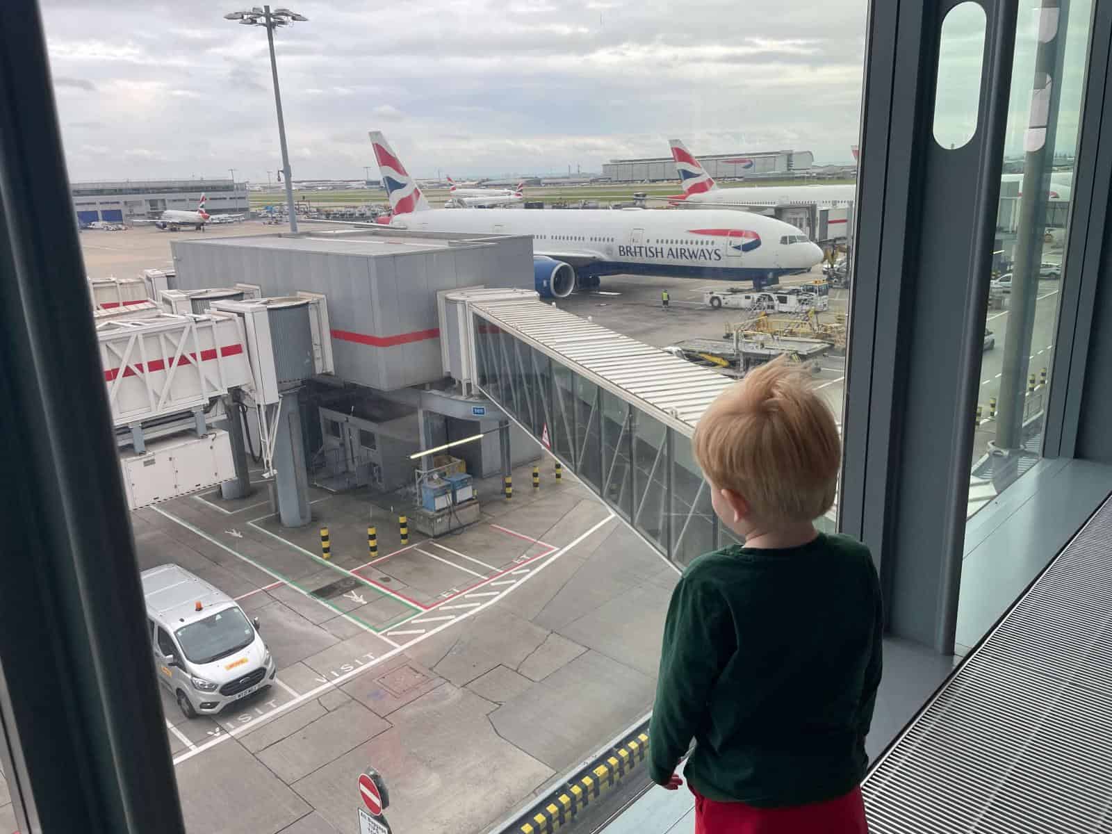 Toddler stood with his back to camera looking through airport window at plane