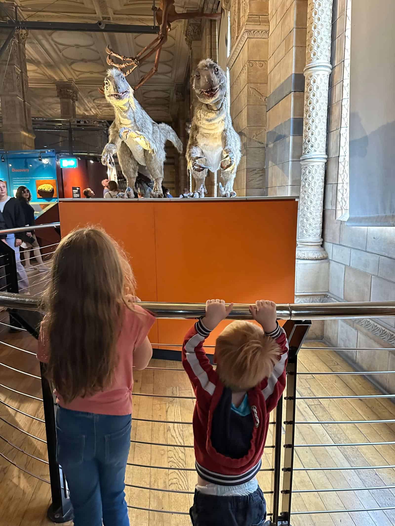 Two children (older girl and toddler boy) looking at two animatronic dinosaurs at the natural history museum in London